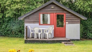 A small wooden cabin with a red door and a window, surrounded by a grassy area with white plastic chairs and orange flowers. - 4 Person Holiday Park Home in Grindsted (Grindsted)