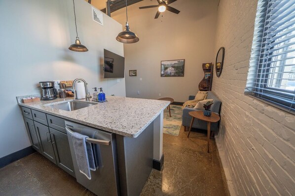 A compact kitchen with a central island, stainless steel appliances, pendant lighting, and a view of a living area with a sofa and a small table.