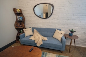 A living room with a blue sofa, a round coffee table, a wooden side table with a plant, and a mirror on the wall.