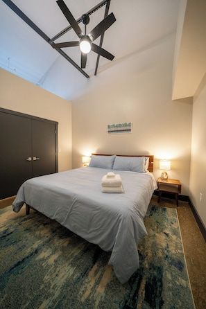 Une chambre à coucher avec un lit, des lampes de chevet, un ventilateur de plafond, une armoire sombre et un tableau au mur.