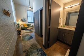 A modern interior with a brick wall, a green sofa, a round mirror, and a kitchen area with cabinets and a countertop.