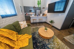 A compact living space with a kitchenette, a green sofa, a yellow throw blanket, and a round wooden coffee table.