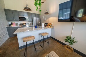 A modern kitchen with a central island, bar stools, a large TV, and hanging plants.