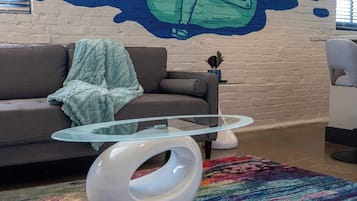 A modern living room with a glass coffee table, a colorful rug, a sofa with a throw blanket, and a mural on the wall.