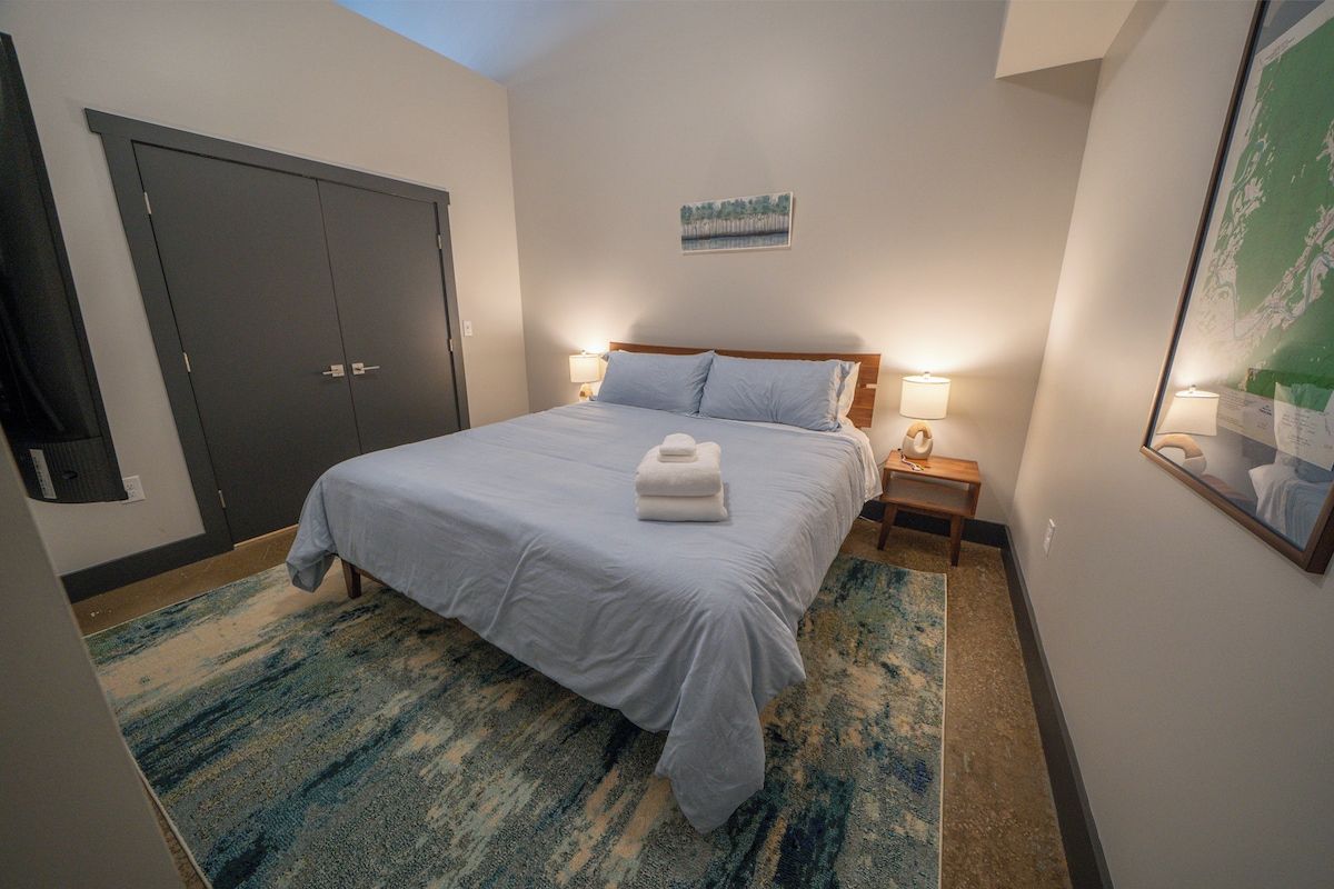 Habitación de hotel con una cama, lámparas de noche, una mesita de noche, una puerta oscura grande y un cuadro en la pared.