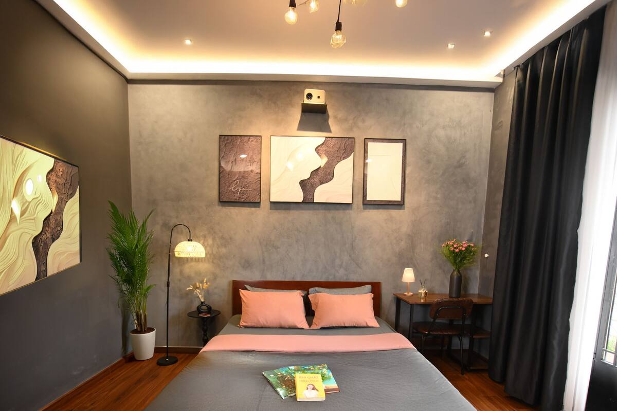 A modern bedroom with a large bed, a bedside table with a lamp, a small table with a vase, and a potted plant.