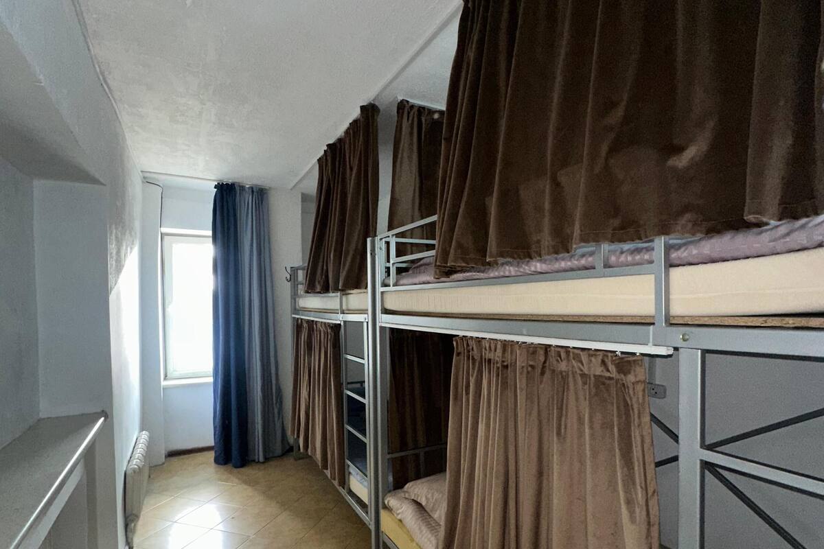 A dormitory room with bunk beds, a radiator, a window with curtains, and a tiled floor.