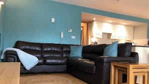 A living room with a dark leather sofa, a small wooden table, and a kitchen in the background. - Marina Waterfront Apartment - family & pet-friendly near to pubs & beach (Milford Haven)