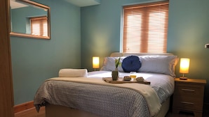 A neatly made bed with a patterned bedspread, a bedside table with a lamp, and a window with blinds. - Marina Waterfront Apartment - family & pet-friendly near to pubs & beach (Milford Haven)