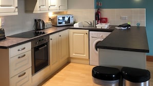A modern kitchen with white cabinets, a microwave, a kettle, a sink, and a washing machine. There are two stainless steel trash bins and a fire blanket on the counter. - Marina Waterfront Apartment - family & pet-friendly near to pubs & beach (Milford Haven)