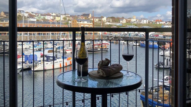 A balcony with a table, a bottle of wine, and a glass of red wine. The balcony overlooks a marina with boats and a town in the background. - Marina Waterfront Apartment - family & pet-friendly near to pubs & beach (Milford Haven)