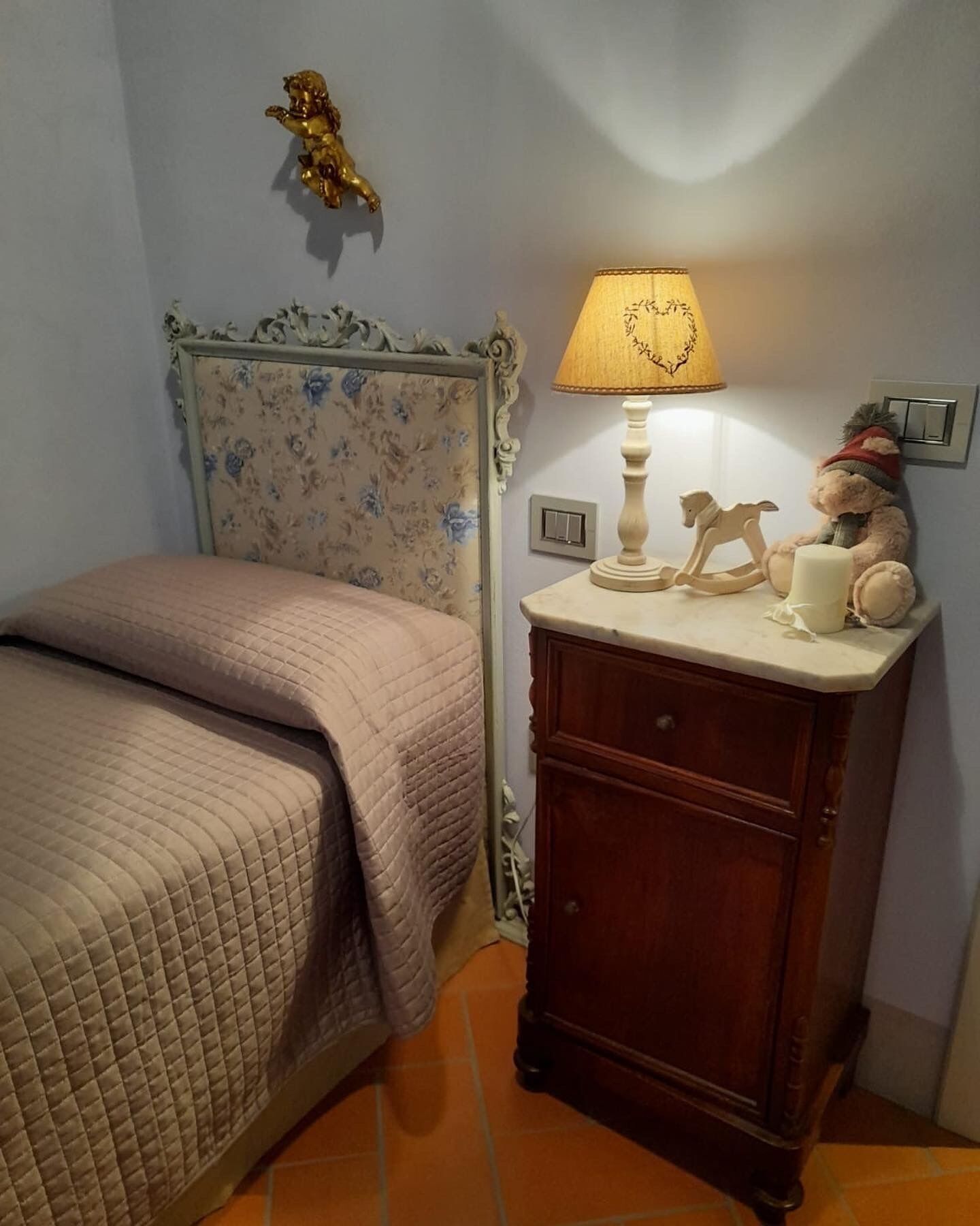 Un dormitorio con cama, una mesita de noche con lámpara, un oso de peluche y un ángel decorativo en la pared.