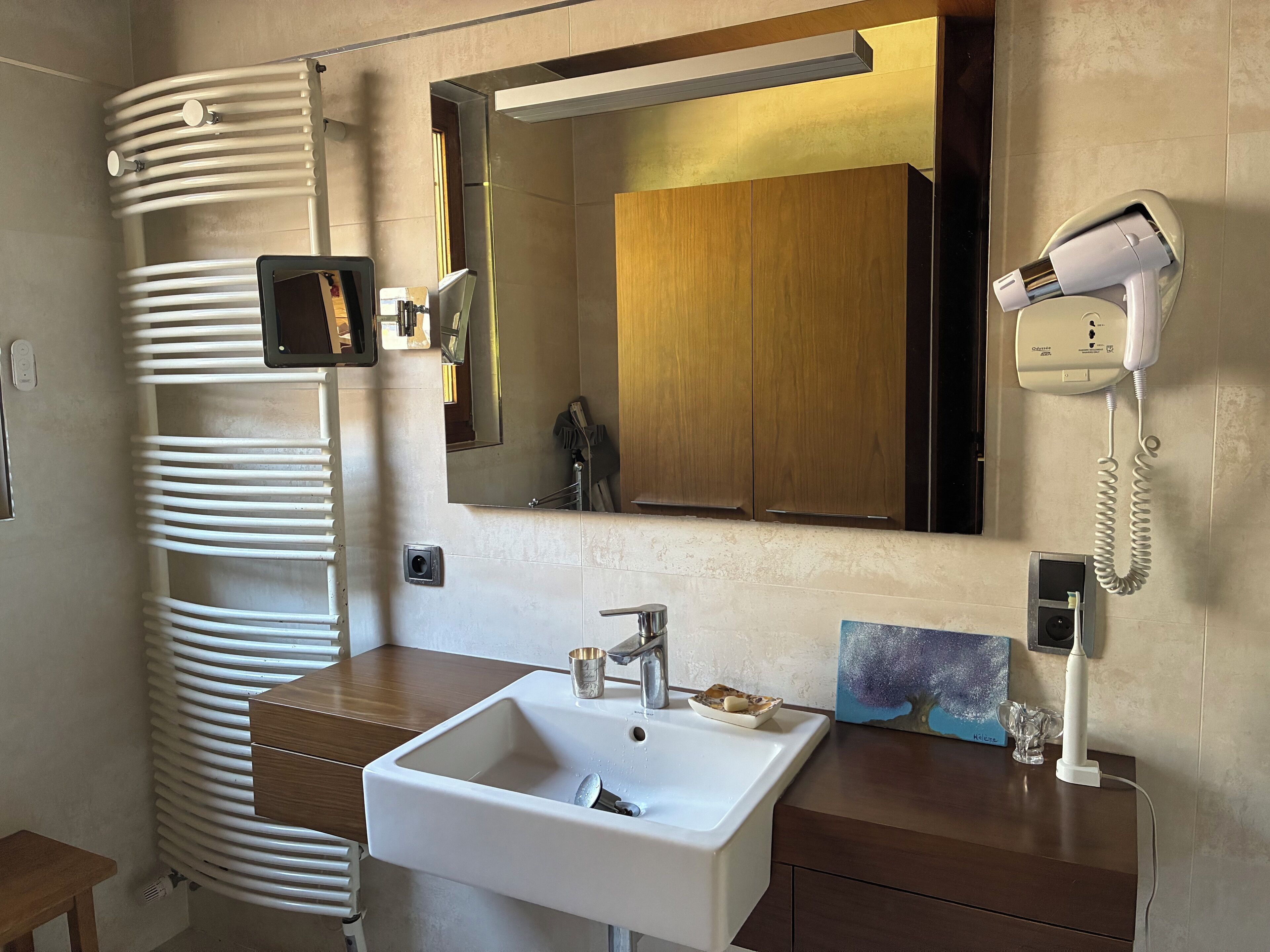Une salle de bain avec un lavabo blanc, un meuble-lavabo en bois, un miroir, un sèche-cheveux et un sèche-serviettes.