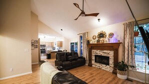A spacious living room with a fireplace, a ceiling fan, and a view of the kitchen. - Heated Swim Spa + Outdoor Movie Theater, Private Resort (Kansas City)