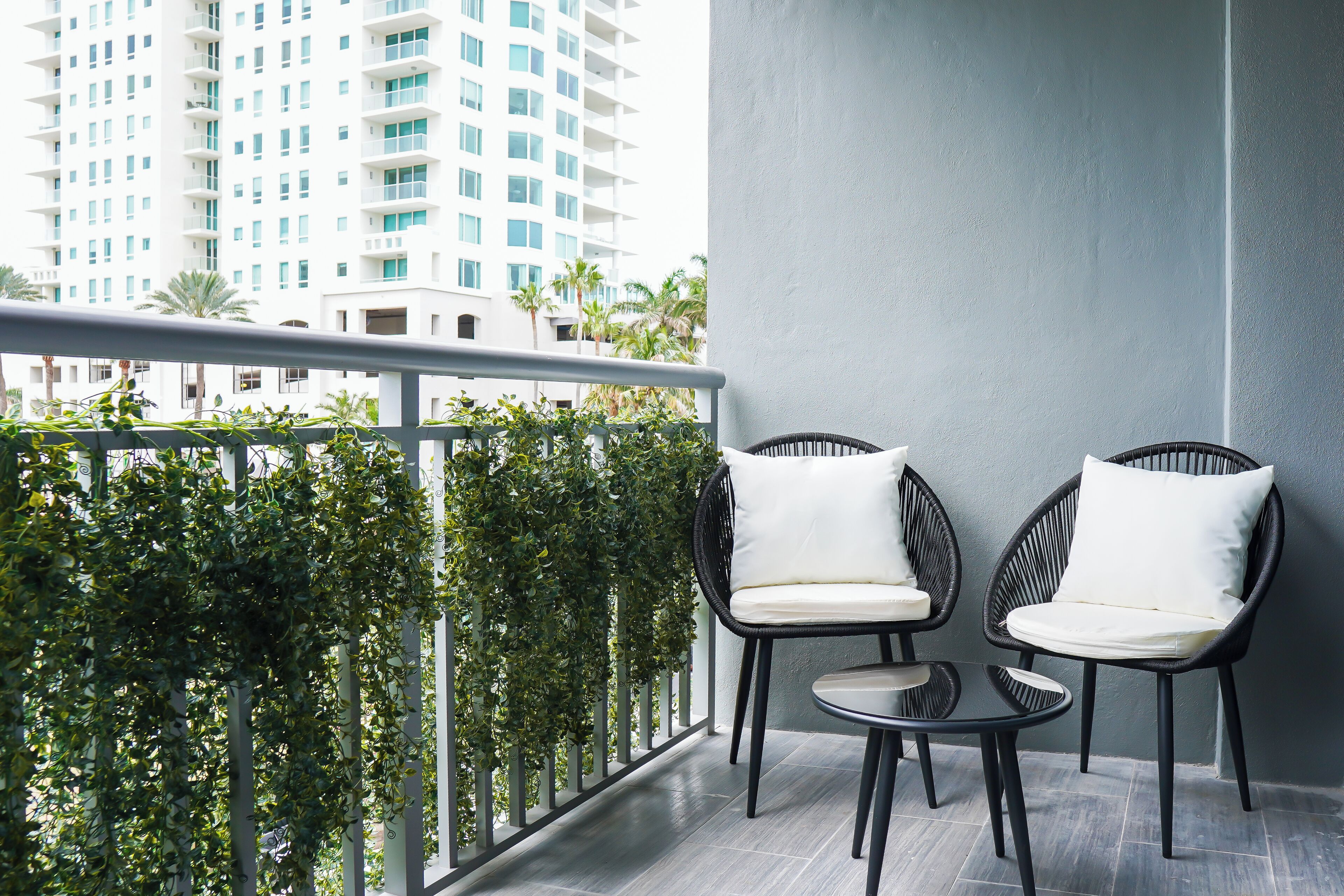 A balcony with two wicker chairs and a small table, surrounded by greenery and overlooking a cityscape.