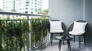 Ein Balkon mit zwei Korbstühlen und einem kleinen Tisch, umgeben von Grünpflanzen und mit Blick auf eine Stadtlandschaft.