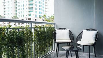 A balcony with two wicker chairs and a small table, surrounded by greenery and overlooking a cityscape.