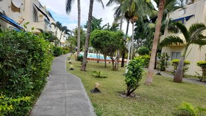 Un chemin mène à un espace piscine entouré de palmiers et d’une pelouse bien entretenue.