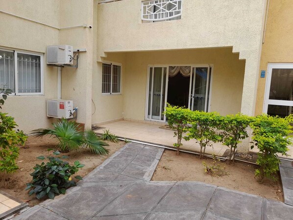 A building with a covered patio area, air conditioning units, and a small garden with various plants.