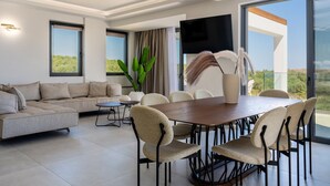 A modern living room with a sofa, dining table, chairs, a TV, and a large window. - Skyline Villa II, By Hellocrete (Aggeliana)