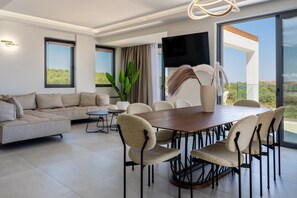 A modern living room with a sofa, dining table, chairs, a TV, and a large window. - Skyline Villa II, By Hellocrete (Aggeliana)