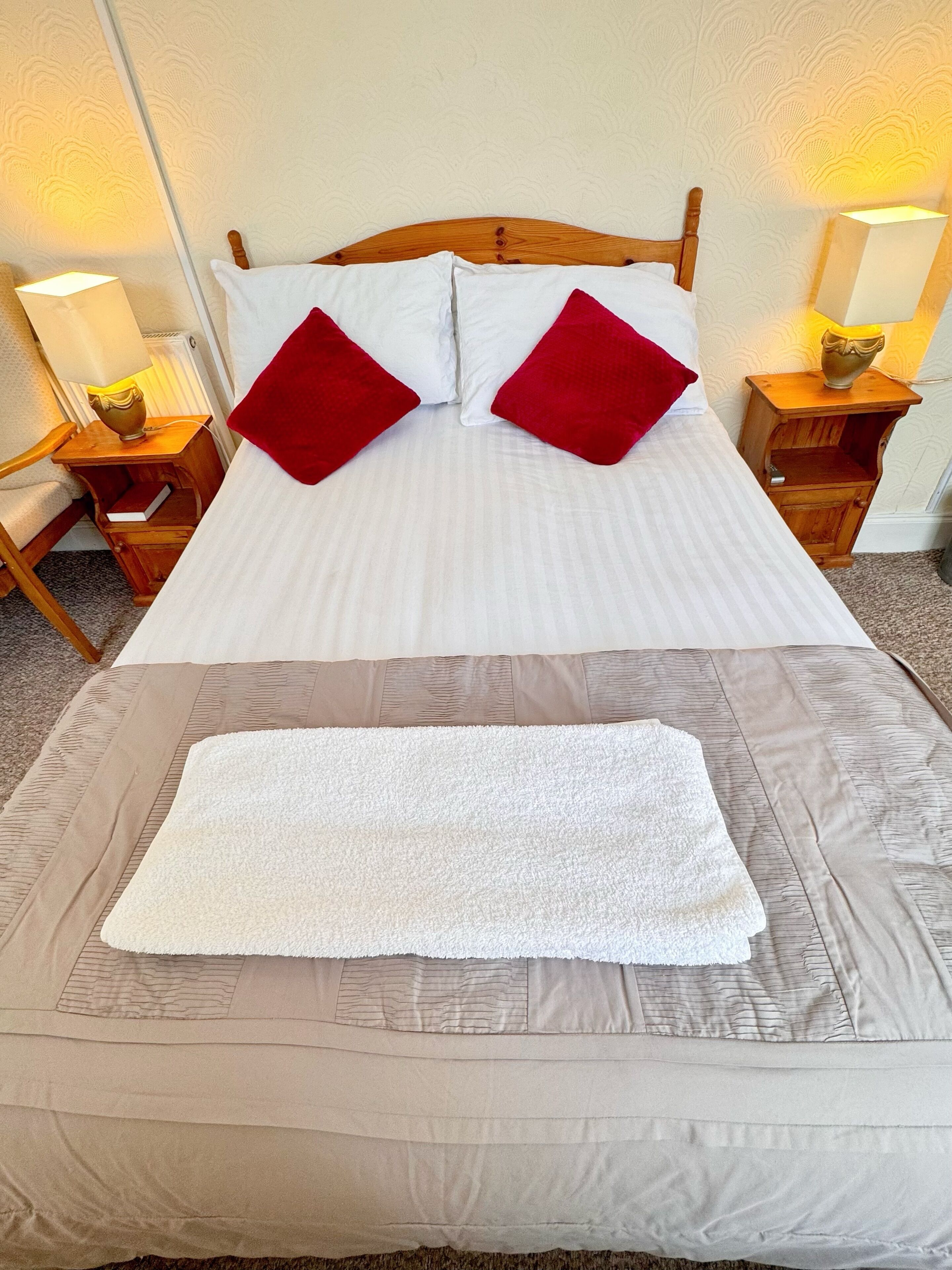 A neatly made bed with white bedding, two red pillows, and a white towel placed on a beige bed runner.