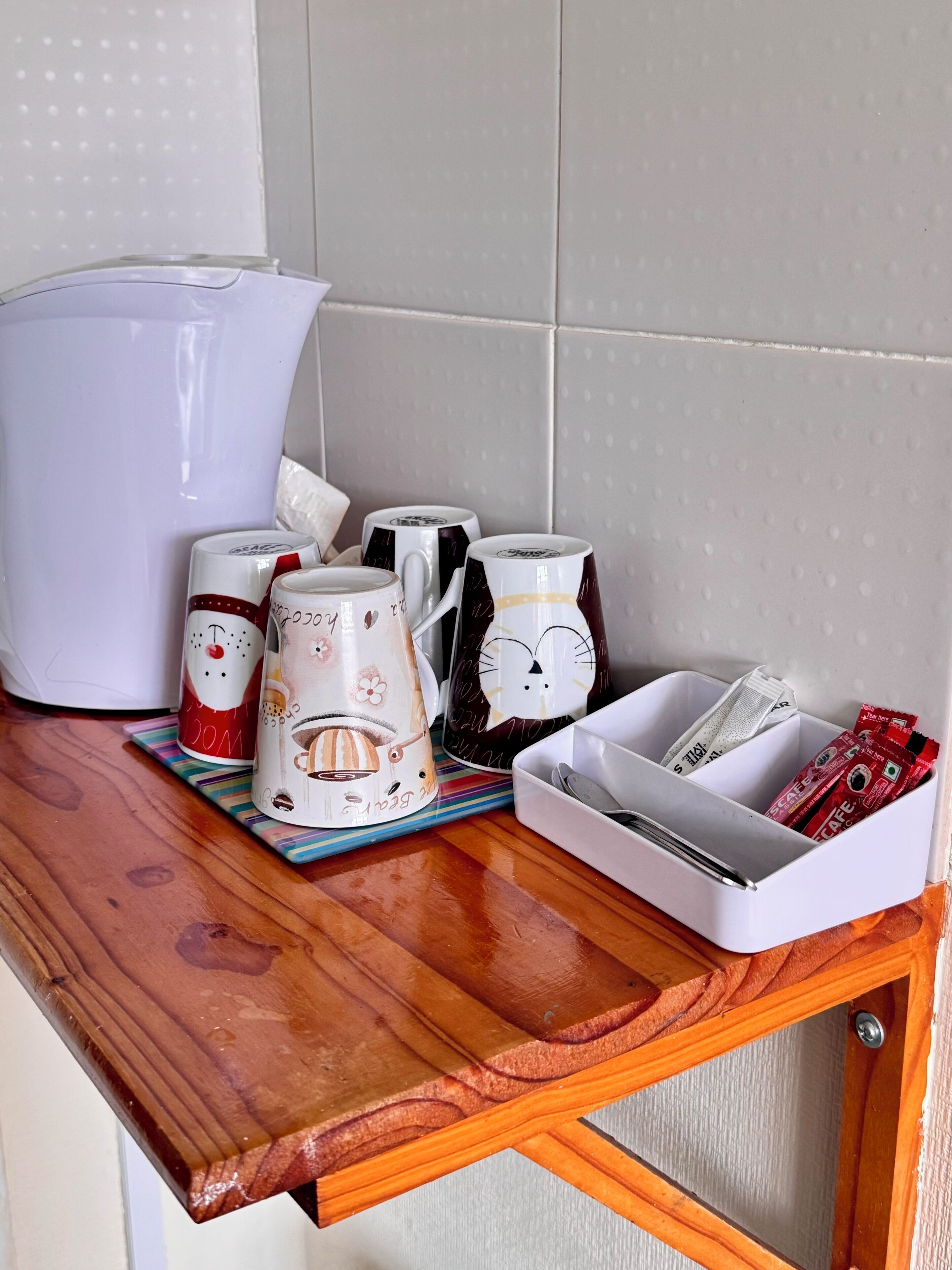 A kitchen counter with various coffee mugs, a white kettle, and a tray with coffee pods and a spoon.