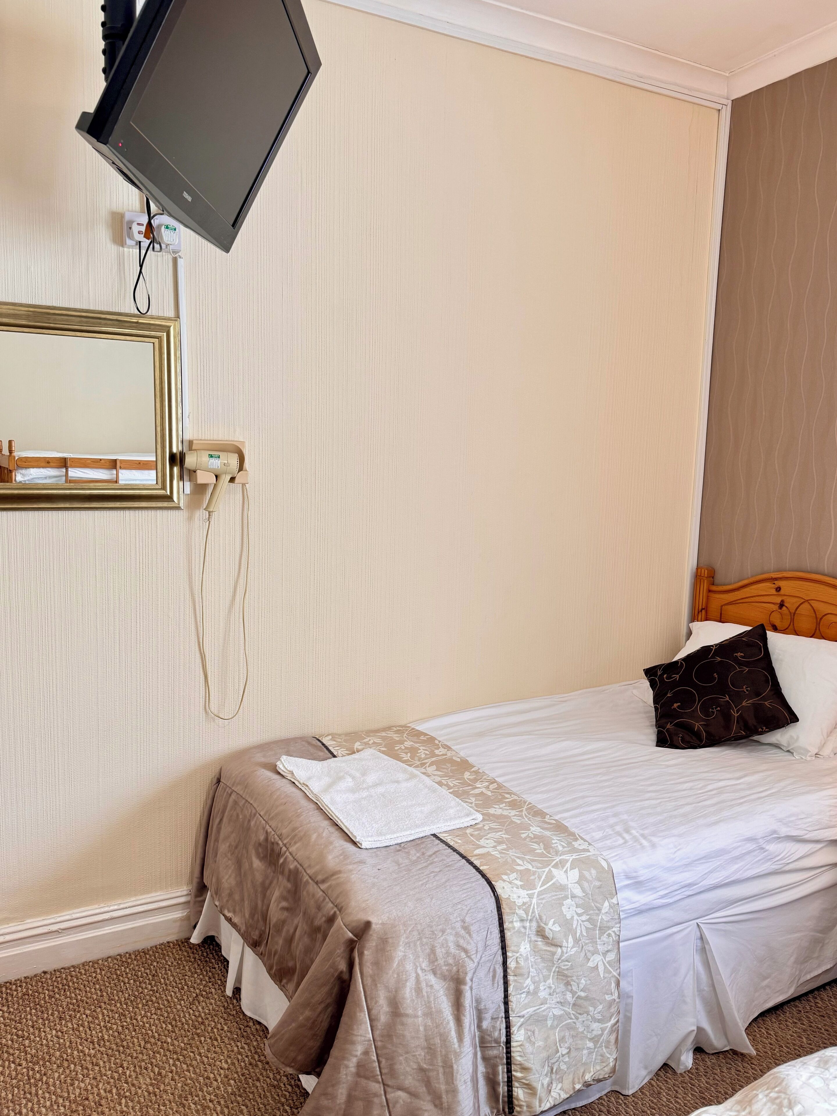 Hotellihuone, jossa on sänky, seinälle asennettu televisio, hiustenkuivaaja ja peili.
