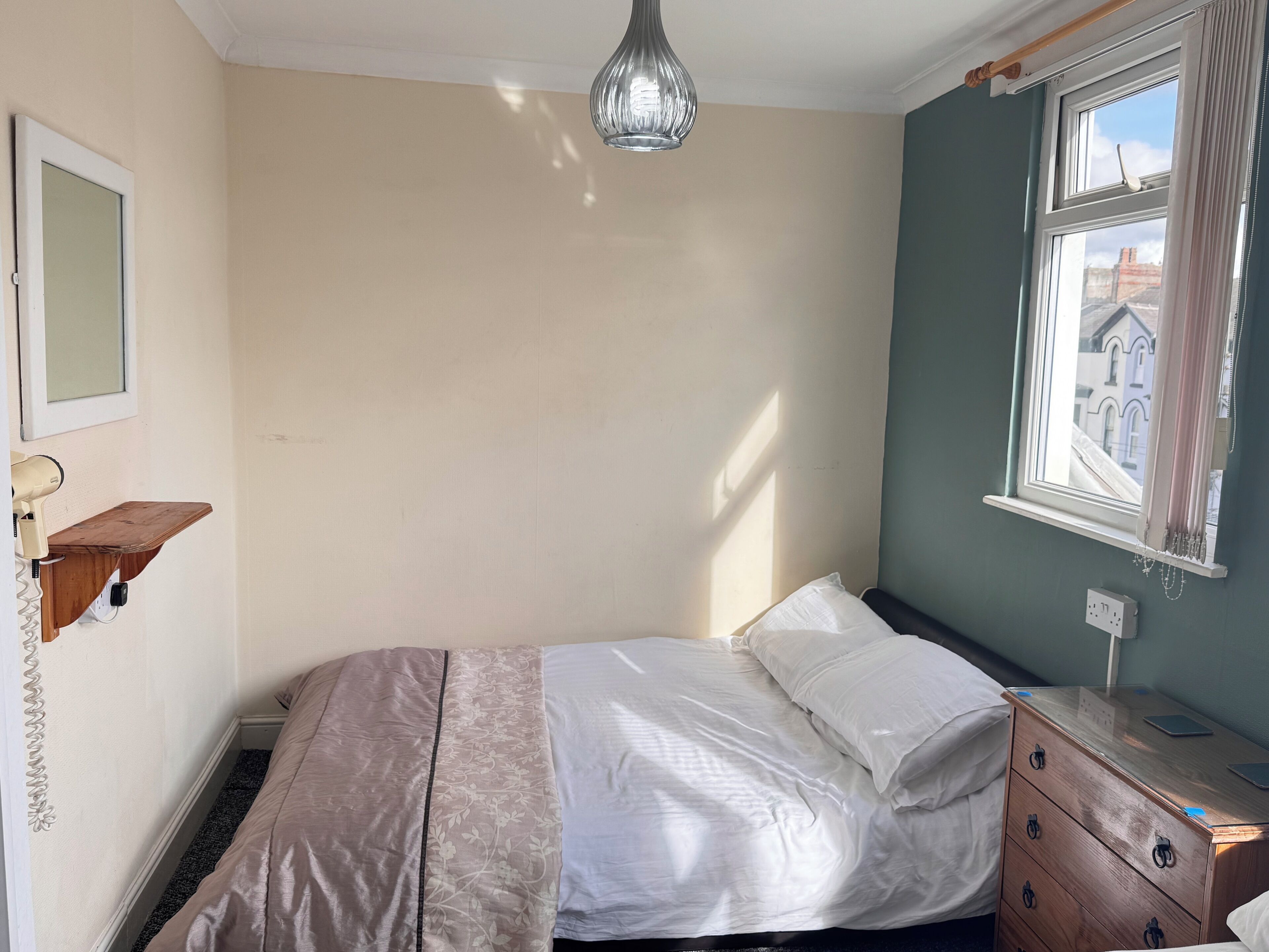 A bedroom with a bed, a dresser, a mirror, a shelf with a hairdryer, and a window with a view of buildings.