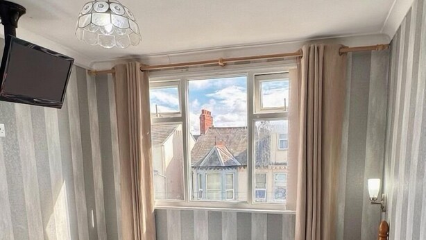 A neatly made bed with white and brown pillows, a wooden headboard, and a window with curtains. - Kendal Hotel (Blackpool)