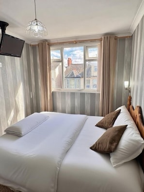 A neatly made bed with white and brown pillows, a wooden headboard, and a window with curtains. - Kendal Hotel (Blackpool)
