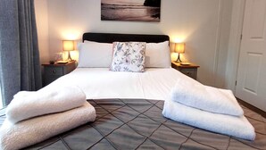Un lit bien fait, avec une tête de lit matelassée, deux lampes de chevet, un coussin à motifs floraux et deux serviettes posées sur le lit.