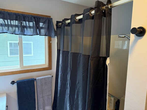 Un baño con cortina de ducha, una ventana con persianas y dos toallas colgadas en un perchero.