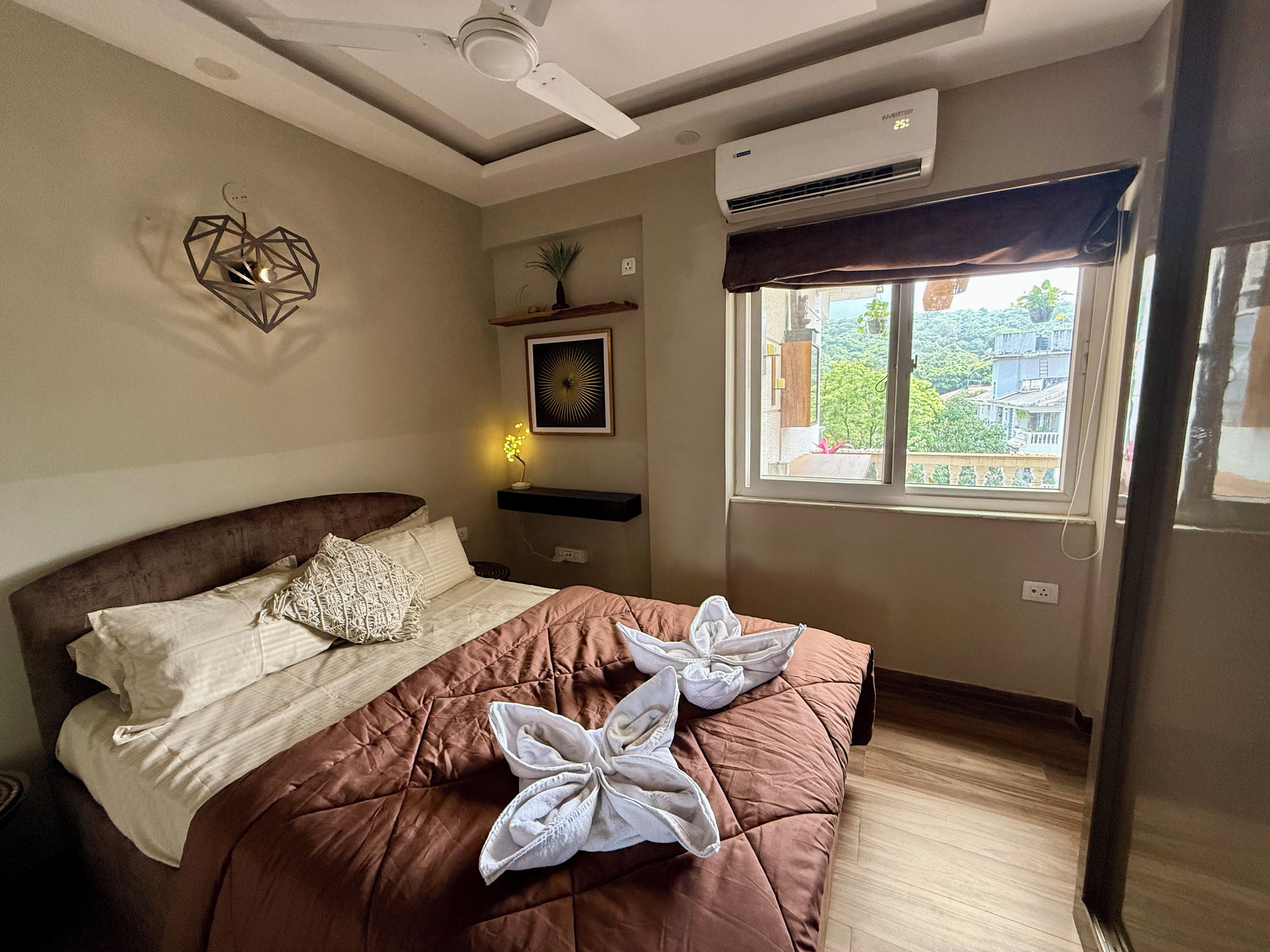 Una camera d'albergo con un letto, una ventola a soffitto, un condizionatore d'aria, una finestra con vista e una mensola a parete con una pianta.