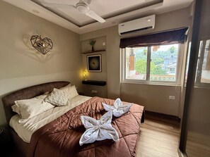 A hotel room with a bed, a ceiling fan, an air conditioning unit, a window with a view, and a wall-mounted shelf with a plant. - Dishney Den- 1BHK - Hill View - Pvt Balcony Bar (Shivoli)