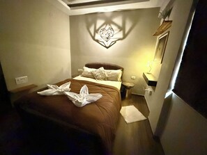 Una cama bien tendida con una colcha marrón y almohadas blancas, una mesita de noche con una lámpara, y un aplique decorativo en la pared.