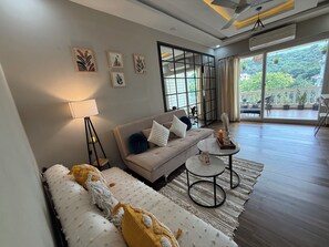 A modern living room with a sofa, a coffee table, and a lamp. There is a balcony with potted plants and a view of greenery. - Dishney Den- 1BHK - Hill View - Pvt Balcony Bar (Shivoli)