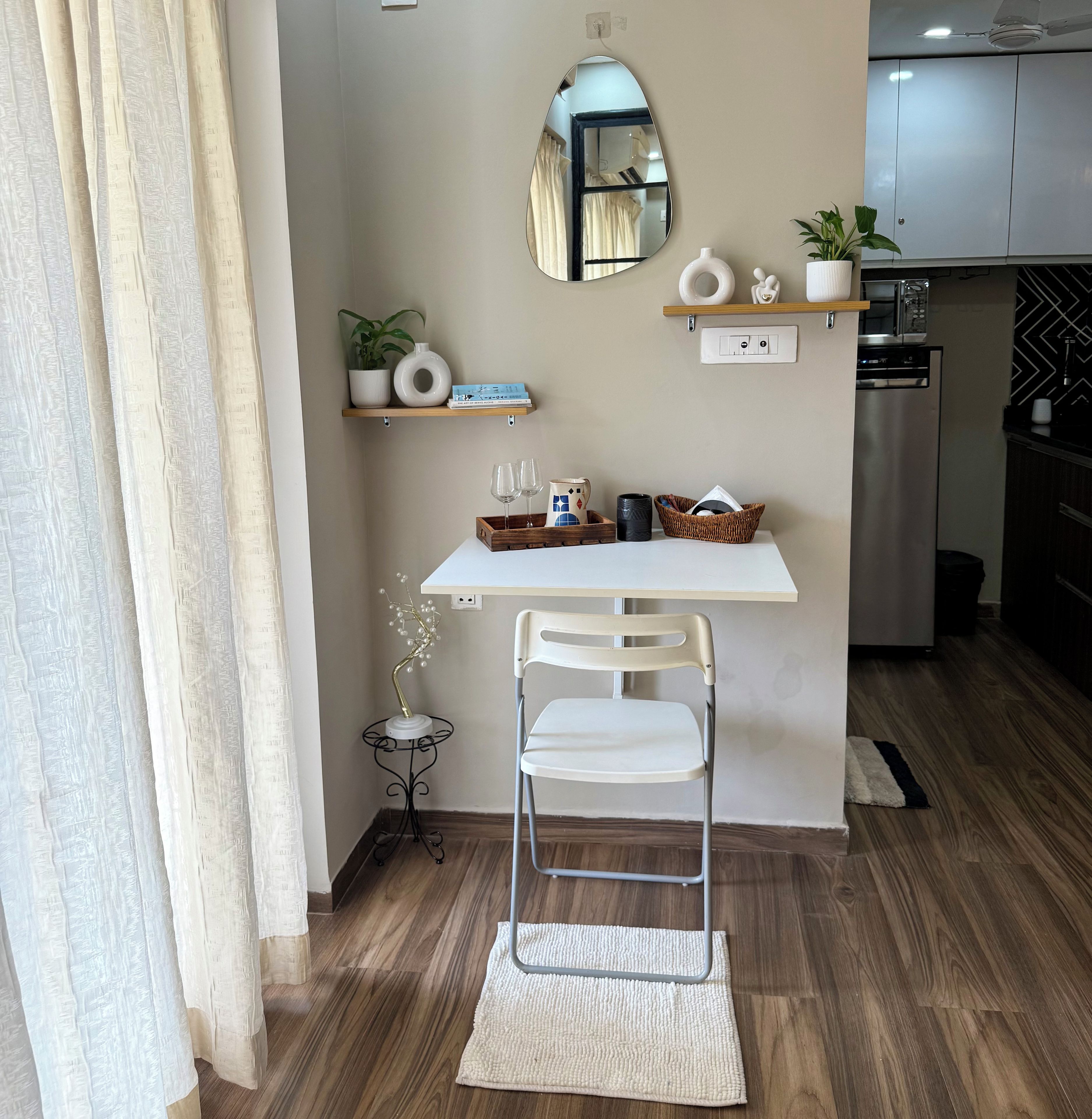 Una kitchenette compacta con encimera blanca, una pequeña zona de comedor con una silla blanca, un espejo y una planta en maceta.