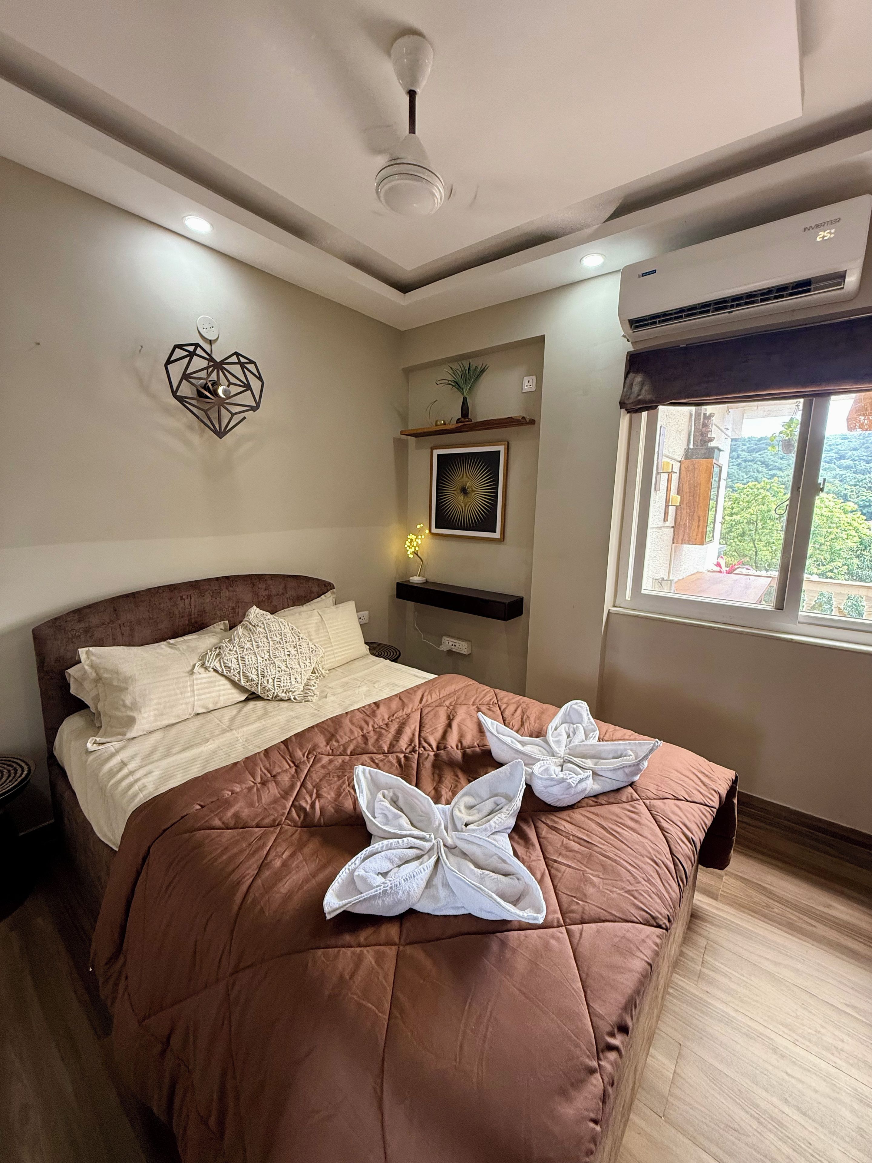 A bedroom with a bed, a ceiling fan, an air conditioning unit, a wall-mounted shelf with a plant, and a framed picture.