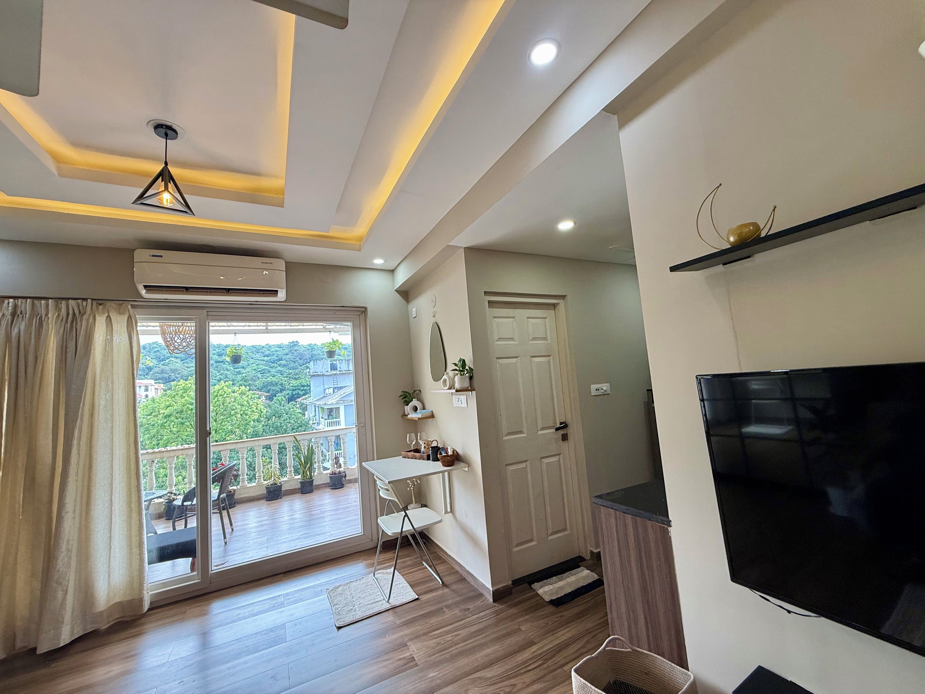A modern living room with a large TV, a balcony with potted plants, and a sliding glass door.