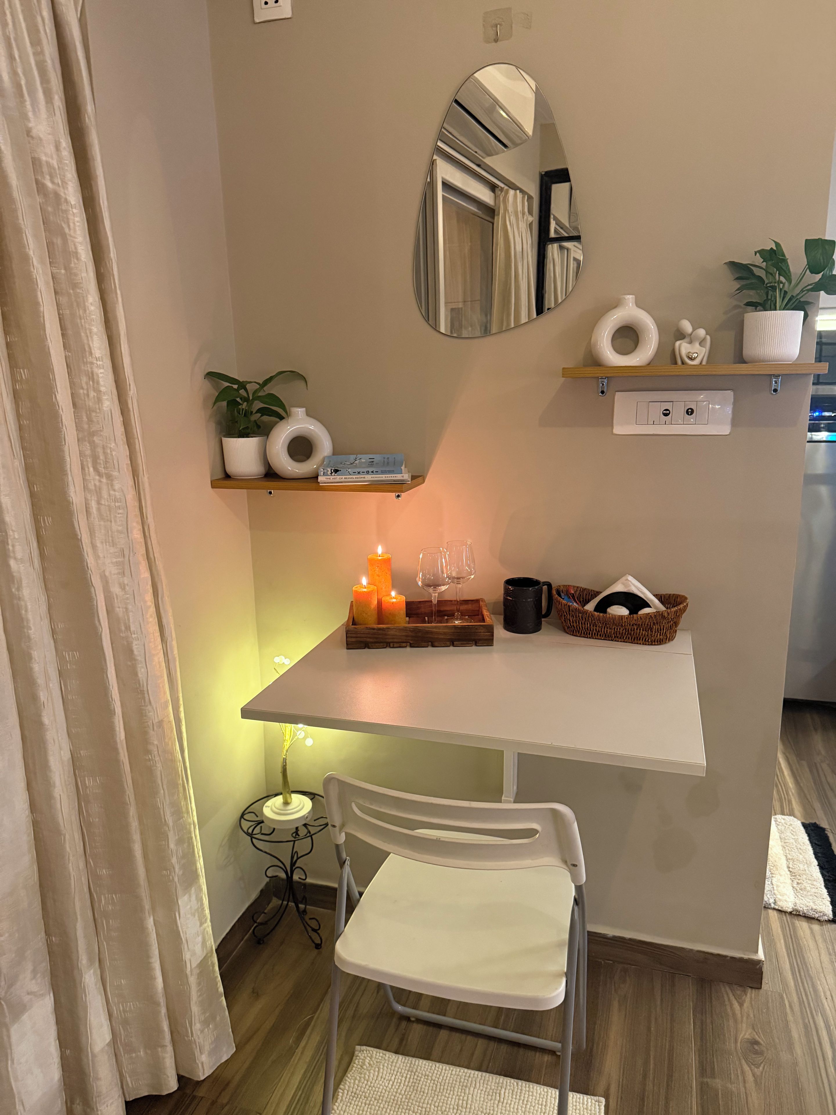 Habitación pequeña con una mesa blanca, una silla, un espejo, una estantería con adornos y una mesita de noche con una vela.