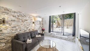 Een moderne woonkamer met een stenen muur, een grijze bank, een glazen salontafel en uitzicht op het groen door grote ramen.