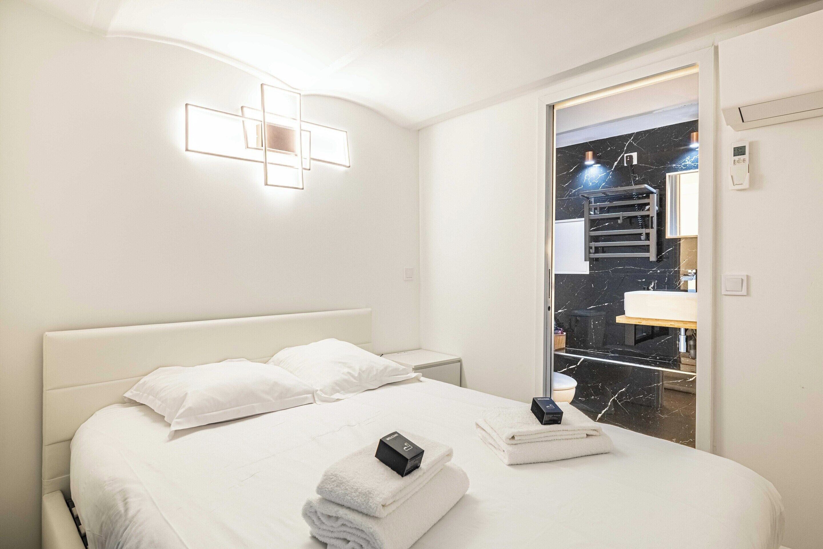 A hotel room with a bed, white linens, a bedside table, a nightstand, a modern light fixture, and a bathroom visible through a glass door.