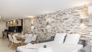 Een moderne hotelkamer met een stenen muur, een bed met witte lakens, een zithoek met een tafel en stoelen, en een kitchenette op de achtergrond.