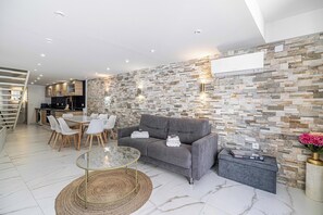 A modern living room with a grey sofa, a glass coffee table, and a dining area with white chairs.