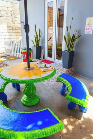 A patio area with a round table, two blue and green stools, a potted plant, and a sign on the wall.