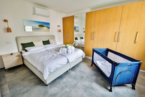 A modern bedroom with a large bed, a wooden wardrobe, a baby crib, and a wall-mounted air conditioner.