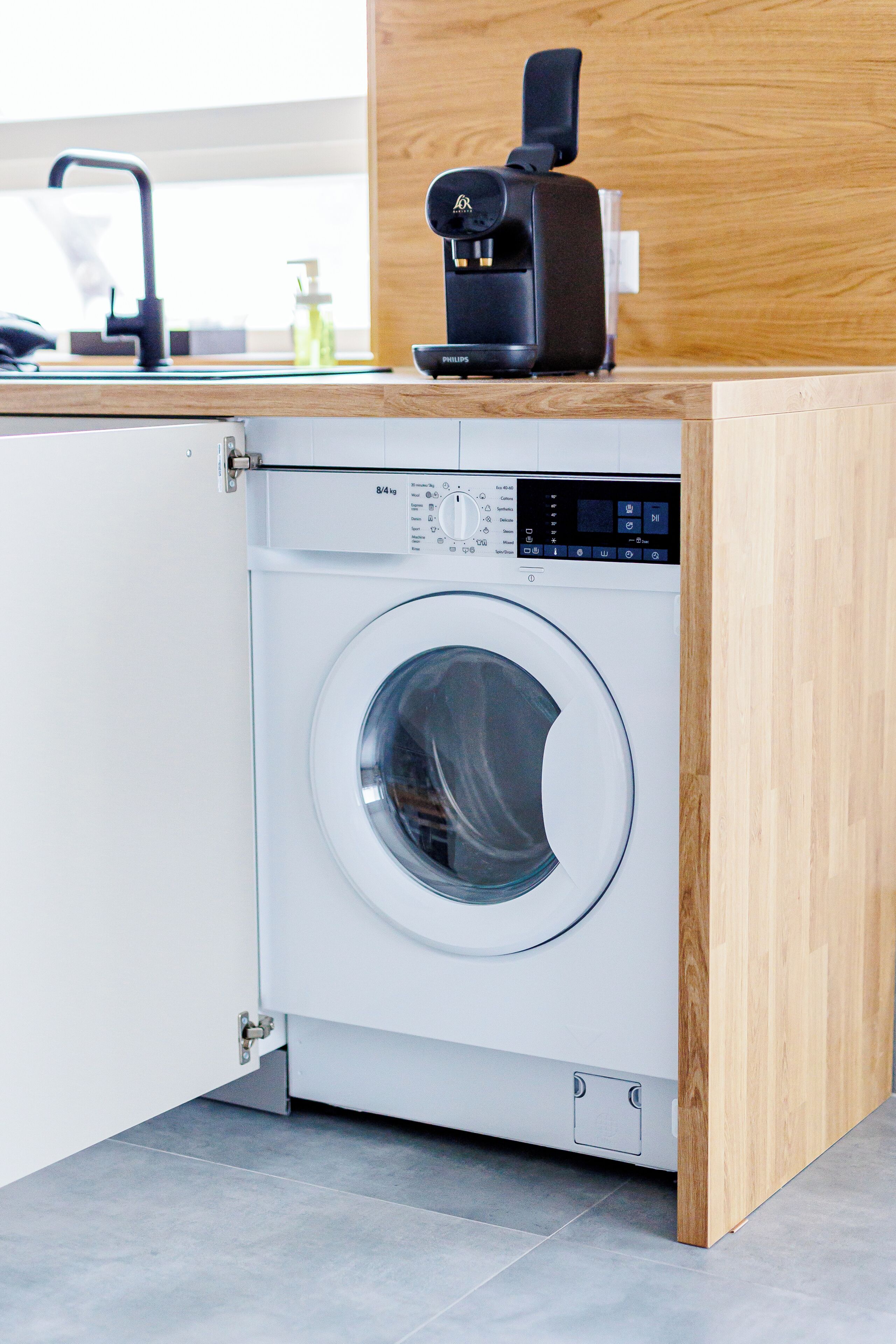 Eine kompakte, moderne Waschmaschine, integriert in eine Holz-Arbeitsplatte, mit einer digitalen Bedienblende und einer transparenten Tür.