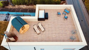 A rooftop terrace with a wooden deck, a blue patterned sofa, two lounge chairs, a small table, and a fan.