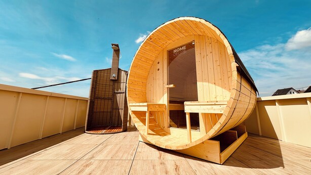 A wooden sauna structure with a door labeled "HOME DELUXE" on a rooftop with wooden flooring.